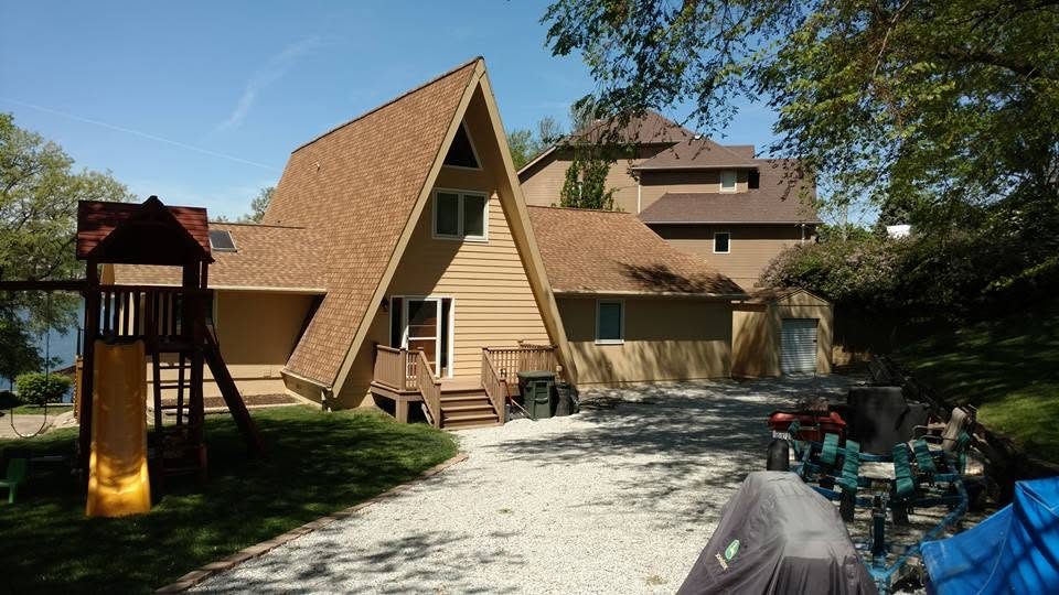 Large A-frame house with tan siding, brown roof, wooden deck, and playground equipment on gravel driveway surrounded by trees