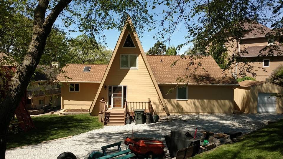Residential A-frame house with cream siding, wooden deck, surrounded by mature trees and landscaping