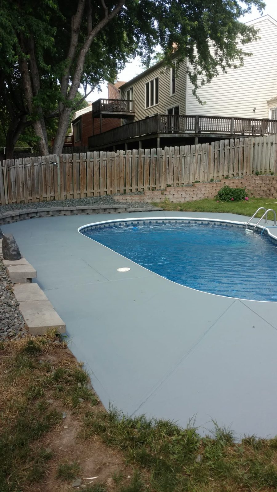 In-ground swimming pool with concrete deck, wooden fence, and residential house with deck in background